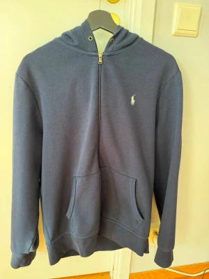 Mörkblå hoodie från Polo Ralph Lauren - Jag säljer en snygg mörkblå hoodie från Polo Ralph med full zip. Denna är i ett mycket bra skick och endast använd ett fåtal gånger, den kom ej till användning. Hör av er vid funderingar!