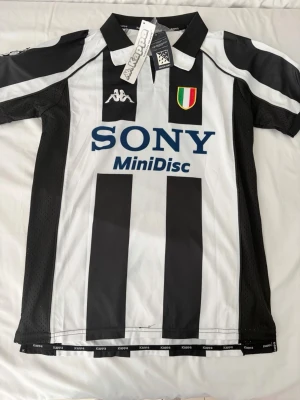 Juventus matchtröja Del Piero Kappa - Juventus klassiska svartvit randig matchtröja från Kappa med Del Piero och nummer 10 på ryggen. Tröjan har SONY MiniDisc som sponsortryck, klubbmärke och italienska mästarmärket på bröstet. Tillverkad i lätt och ventilerande material, perfekt för fotboll.