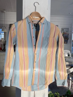 Randig jeansskjorta från Zara i pastell - Säljer en långärmad liknande jeansskjorta från Zara med breda pastellfärgade ränder i ljusblått, beige och vitt samt tunna röda detaljer. Skjortan har klassisk krage med knappar och bröstficka. Perfekt för dig som gillar färg och en avslappnad stil.