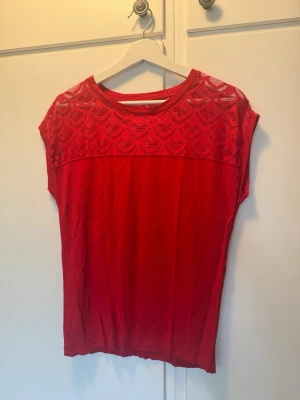 Röd topp med spetsdetalj - Snygg röd topp med ärmlös design och dekorativ spets upptill. Toppen har rund halsringning och ett genombrutet mönster i spetsen över axlar och bröst. Perfekt att styla med jeans eller kjol för en trendig look.