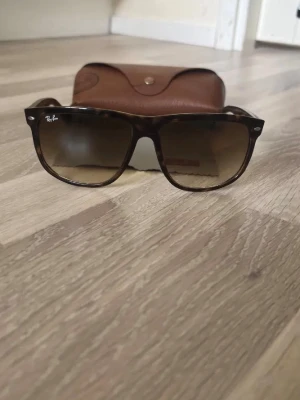 Bruna Wayfarer solglasögon Ray-Ban - Snygga Wayfarer solglasögon från Ray-Ban med brunmelerad båge och mörktonade glas. Klassisk fyrkantig form och diskret Ray-Ban-logga på linsen och skalmen. Perfekta för soliga dagar och kommer med originalfodral.