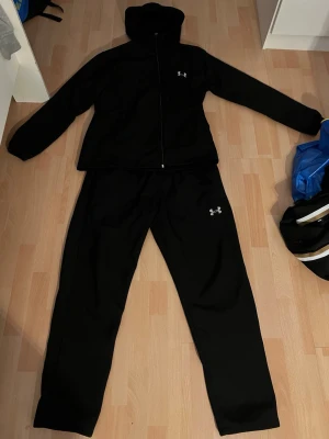 Svart träningsset från Under Armour - Säljer ett svart träningsset från Under Armour med zip-hoodie och matchande byxor. Setet har diskreta loggor på både jacka och byxa, samt en sportig och bekväm passform. Perfekt för gymmet eller löprundan.