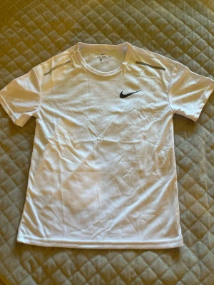 Vit Nike t-shirt i funktionsmaterial - Säljer en vit kortärmad t-shirt från Nike med svart logga på bröstet. Tillverkad i ett lätt och ventilerande funktionsmaterial, perfekt för träning eller sport. Klassisk passform och rund halsringning.
