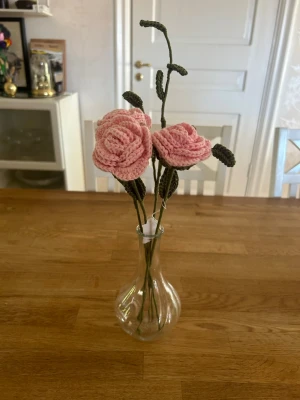 Virkade rosor i vas - Unika handgjorda virkade rosor i ljusrosa garn med gröna stjälkar och blad. Blommorna står i en enkel, genomskinlig glasvas. Perfekt som dekoration till hemmet eller som present till någon speciell.