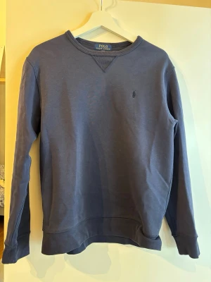 Mörkblå sweatshirt från Polo Ralph Lauren - Klassisk mörkblå sweatshirt från Polo Ralph Lauren med rund halsringning och diskret logga broderad på bröstet. Tillverkad i mjuk bomullsblandning med 41% polyester för extra komfort. Perfekt till jeans eller chinos.