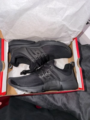  Nike Prestos - Snygga helsvarta Nike Presto sneakers med mesh-ovandel och plastdetaljer vid snörningen. Skorna har en låg siluett och en bekväm, flexibel sula som passar perfekt till streetwear-looken. Klassisk Nike-logga på sidan och tå. Helt oanvända 