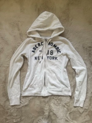 Vit zip up hoodie  Abercrombie fitch - Säljer en vit hoodie med dragkedja från Abercrombie. Tröjan har huva med dragsko, två fickor framtill och mörkblå text med 'Abercrombie 18 New York' broderat på bröstet