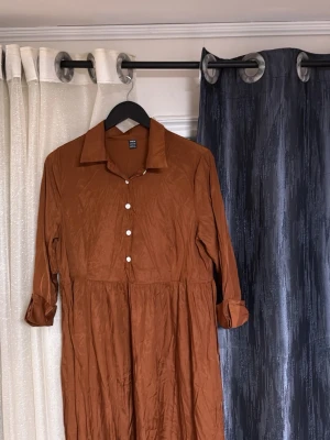 Brun skjortklänning med knappar - Snygg brun skjortklänning med krage och vita knappar framtill. Klänningen har lång ärm som kan vikas upp och en avslappnad passform. Perfekt för dig som gillar enkel men stilren design.