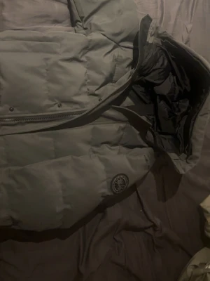 Mörkgrå dunjacka med huva - Snygg mörkgrå dunjacka med stor huva och quiltad design. Jackan har dragkedja framtill, tryckknappar och en rund patch på bröstet. Insidan har svarta detaljer och praktiska innerfickor. Perfekt för kalla vinterdagar.