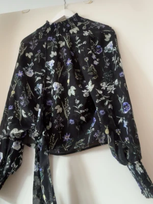 Svart blommig blus med lång ärm - Snygg svart blus med blommönster i lila, vitt och grönt. Blusen har långärmade puffärmar med resår vid handlederna och hög krage med rynk. Tillverkad i ett tunt, lätt material som ger en luftig känsla. Perfekt för att lyfta vilken outfit som helst.
