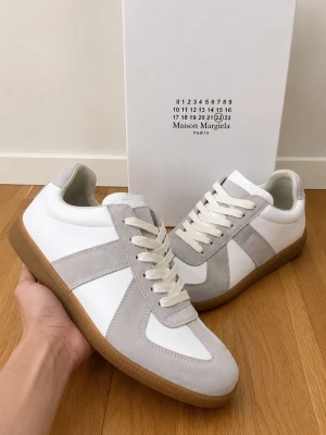 Maison Margiela GATs - Maison Margiela sneakers i vitt och ljusgrått med ovandel i läder och mocka. Klassisk låg modell med vita snören och gummisula. Diskret Maison Margiela-logga på plösen. Perfekta för en clean och stilren look.