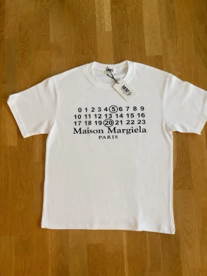 Vit Maison Margiela t-shirt - Säljer en vit t-shirt från Maison Margiela med det ikoniska siffertrycket framtill och rund hals. T-shirten har korta ärmar och är gjord i mjuk bomull. Perfekt för dig som gillar minimalistisk och stilren design.