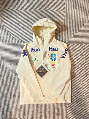 Jordan Brasil vindjacka beige M - Snygg beige vindjacka från Jordan med Brasil-logga och flera sponsortryck. Jackan har huva, hel dragkedja och är tillverkad i GORE-TEX-material som står emot regn och vind. Perfekt för dig som gillar sportig stil och vill sticka ut.