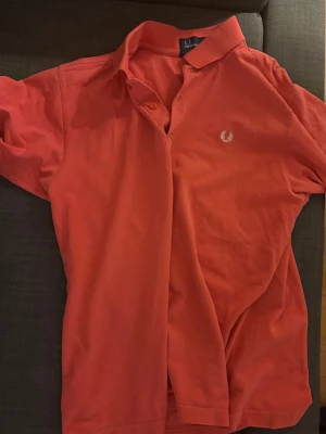 Rosa pikétröja från Fred Perry - Säljer en klassisk röd pikétröja från Fred Perry i 100% bomull. Tröjan har korta ärmar, krage och knappar framtill samt den ikoniska Fred Perry-loggan broderad på bröstet. Perfekt för en avslappnad och stilren look.