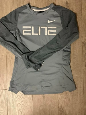 Grå långärmad Nike Elite tröja - Snygg grå långärmad träningströja från Nike med texten ELITE framtill och vit Nike-logga. Tillverkad i funktionsmaterial som andas, perfekt för träning eller sport. Tröjan har raglanärmar och en sportig passform.