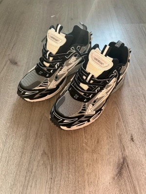 Off-White Be right back svarta och vita sneakers - Säljer ett par Off-White sneakers i svart, vitt och grått med coola grafiska detaljer. Skorna har meshpaneler för bra ventilation och robust sula för extra komfort. Snygga snörningar och Off-White-logga på plösen. Perfekta för dig som gillar streetwear.