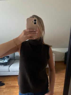 Brun ärmlös polotopp - Mörkbrun ärmlös polotopp med hög krage. Toppen har en loose passform och är tillverkad i ett mjukt stickat material som känns skönt mot huden. Perfekt att styla med jeans eller kjol för en enkel men snygg look.