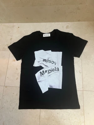 Svart Maison Margiela t-shirt - Cool svart t-shirt från Maison Margiela med ett unikt grafiskt tryck framtill i vitt och svart. Klassisk rund hals och korta ärmar. Perfekt för dig som gillar streetwear och vill sticka ut med en statement tee.