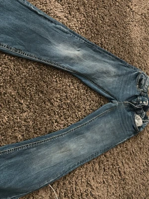 Blå bootcut jeans med slitningar - Säljer ett par blå bootcut jeans med snygga slitningar och klassisk femficksdesign. Jeansen har normalhög midja och stängs med knappar. Perfekta för dig som gillar en avslappnad och trendig stil.
