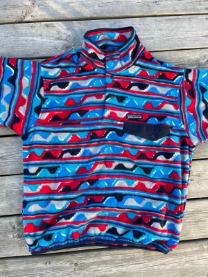 Färgglad fleece från Patagonia - Säljer en mönstrad fleece från Patagonia med färger i rött, blått, svart och vitt. Tröjan har hög krage, tryckknappar och en bröstficka med knapp. Perfekt för dig som gillar retrostil och vill sticka ut med ett unikt plagg.