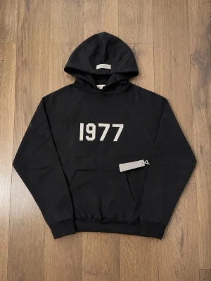1977 iron grey essentials hoodie (XXS-JUNIOR) - Snygg svart hoodie med stor huva och trycket '1977' i vitt på bröstet. Hoodien har en klassisk känguruficka framtill och ribbade muddar vid ärmar och nederkant. Perfekt för en avslappnad och trendig look. XXS JUNIOR PASSFORM 164/5-177. Oversized ish passform