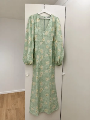 Grön blommig långklänning från Adoore - Ljusgrön långklänning från Adoore med v-ringning, långa puffärmar och blommigt mönster i vitt och gult. Klänningen har en smickrande passform och dragkedja i ryggen. Perfekt för sommarens alla tillfällen.