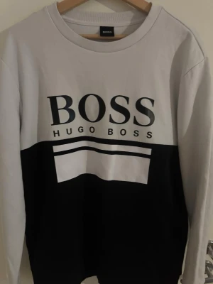 Svartvit sweatshirt från Hugo Boss - Snygg svartvit sweatshirt från Hugo Boss med stor logga på bröstet. Tröjan har rund halsringning och långa ärmar. Perfekt för dig som gillar stilrena och klassiska plagg med tydlig branding.