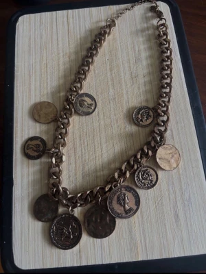 Kedjehalsband med medaljonger från P&B samt ett halsband med matchande öronhänge - Unikt halsband i antikguldfärgad metall från P&B. Kedjan är chunky och pryds av flera medaljonger med olika motiv, bland annat profiler och djur. Perfekt för dig som gillar statement-smycken och vintagevibbar. Lås med karbinhake och justerbar längd.  Sen har vi ett halsband med matchande öronhänge i rostfritt stål.