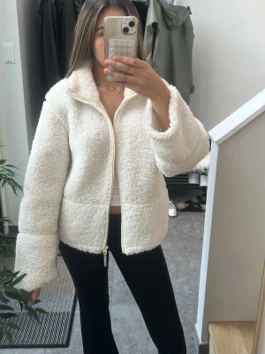 Vit teddyjacka med dragkedja - Mysig vit teddyjacka i fluffigt material med hög krage och dragkedja framtill. Jackan har en avslappnad passform och långa ärmar, perfekt för att hålla värmen under kyliga dagar. Enkel och stilren design som passar till det mesta. Originalpris 400