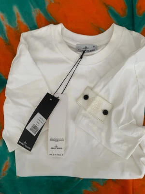 Vit Stone Island t-shirt med patch - Säljer en clean vit t-shirt från Stone Island med den klassiska patchen på ärmen. T-shirten har rund hals och korta ärmar, tillverkad i mjuk bomull. Perfekt för dig som gillar stilrena plagg med ikoniska detaljer.