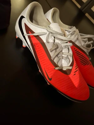 Nike fotbollsskor röd och vit - Snygga fotbollsskor från Nike i rött, vitt och svart med klassisk swoosh-logga på sidan. Skorna har snörning, låg profil och dobbar för bra grepp på planen. Perfekta för dig som vill sticka ut på matchen och få bra bollkontroll.