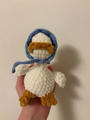 Virkad gås med blå hatt - Söt handgjord gås i mjukt vitt garn med gula fötter och näbb. Gåsen har en blå virkad hatt runt huvudet och svarta ögon. Perfekt som dekoration eller present till någon som gillar gulliga saker.