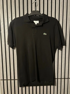 Svart pikétröja från Lacoste - Klassisk svart pikétröja från Lacoste med korta ärmar och krage. Tröjan har den ikoniska gröna krokodilloggan på bröstet och är tillverkad i bomull för en bekväm känsla. Perfekt till jeans eller shorts för en clean look. Använd 2 gånger och pris kan diskuteras (ny pris 1200)