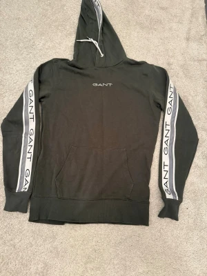 Svart hoodie från GANT med logga - Säljer en snygg mörkgrön hoodie från GANT i storlek S. Tröjan har vita band med GANT-logga längs ärmarna och insidan av huvan, samt en mindre logga på bröstet.  Märke: GANT  Storlek: S (Small)  Färg: Mörkgrön / Khaki  Skick: Mycket gott skick. Inga hål, fläckar eller synligt slitage. Snören och tryck är helt intakta. 