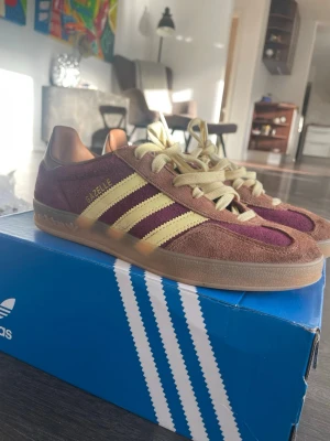 Adidas Gazelle vinröd/gul mocka - Säljer ett par Adidas Gazelle Indoor sneakers i vinröd och gul mocka med klassiska tre ränder på sidan. Skorna har beige snörning och gummisula. Perfekta för dig som gillar retrostil och vill sticka ut med färg. Modellnamnet syns i guld på sidan.