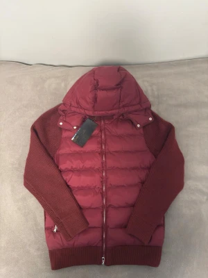 Marcel classic 02 hyberjacka - Snygg vinröd pufferjacka med avtagbar huva och stickade ärmar. Jackan har quiltad front, ribbade muddar och dragkedja framtill. Perfekt för kalla vinterdagar och ger en trendig look med sin kombination av material.