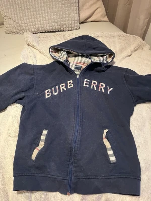 Marinblå Burberry hoodie med dragkedja - Snygg marinblå hoodie från Burberry med dragkedja och huva. Klassisk Burberry-rutig insida på huvan och detaljer vid fickorna. Stor logga i tygbokstäver över bröstet. Perfekt för dig som gillar exklusiv streetwear och vill sticka ut.