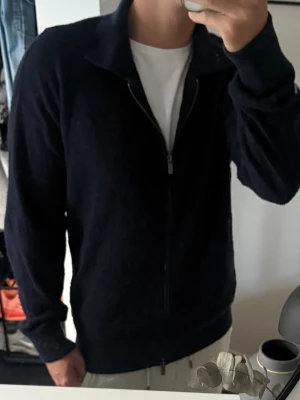 Mörkblå stickad ziptröja från Massimo dutti - Snygg mörkblå stickad tröja med dragkedja framtill och klassisk krage från Massimo dutti. Tröjan har långa ärmar och ribbade muddar vid ärmslut och nederkant. Perfekt att slänga på sig över en t-shirt för en clean look. Ganska nopprig, annars bra skick 