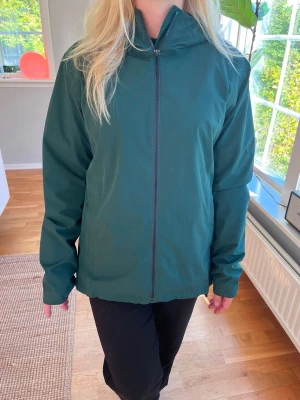 Mörkgrön vindjacka med dragkedja från Salomon - Snygg mörkgrön vindjacka med hel dragkedja framtill och hög krage. Jackan har en minimalistisk design, raka ärmar och sidofickor med dragkedja. Tillverkad i ett lätt och vindtätt material som passar perfekt för blåsiga dagar.