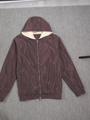 Brun vindjacka med huva - Snygg brun vindjacka med dragkedja och huva. Jackan har en ljus insida, två sidofickor och resår vid ärmslut och midja. Tillverkad i ett lätt, vindtätt material som passar perfekt för blåsiga dagar.