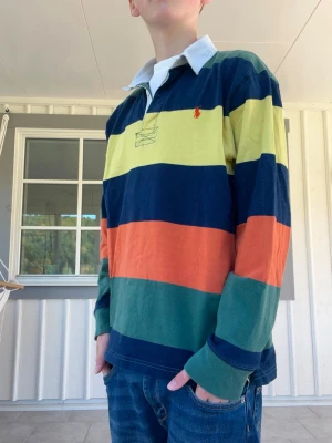 Randig rugbytröja från Polo Ralph Lauren - Säljer en färgglad rugbytröja från Polo Ralph Lauren med breda ränder i marinblått, gult, orange och grönt. Tröjan har vit krage, knappslå och den klassiska röda loggan broderad på bröstet. Perfekt för dig som gillar preppy stil och vill sticka ut. Storlekslappen är borta men skulle tro att den är 14-16