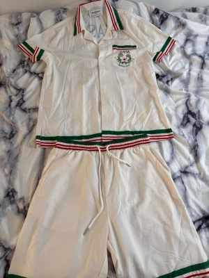 Casablanca set - Snyggt Casablanca set i vitt med gröna och röda ränder på krage, ärmslut, midja och ben. Helt ny oanvänd i storlek S.                                              Skjortan har kort ärm, krage och bröstficka med broderad blomma och texten 'CASA Tennis Club'. Matchande shorts med dragsko i midjan.