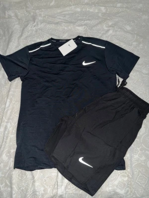 Svart Nike träningsset t-shirt & shorts - Säljer ett svart träningsset från Nike med t-shirt och shorts. Båda plaggen har den klassiska vita Nike-loggan och är tillverkade i ett lätt, snabbtorkande material som passar perfekt för träning. T-shirten har rund hals och reflexdetaljer på axlarna.