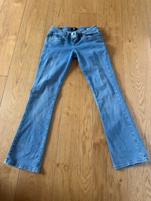 Blå bootcut jeans med låg midja - Säljer ett par ljusblå bootcut jeans med låg midja och klassiska fem fickor. Jeansen har dragkedja och knapp framtill samt utsvängda ben. Perfekta för en avslappnad och trendig look.