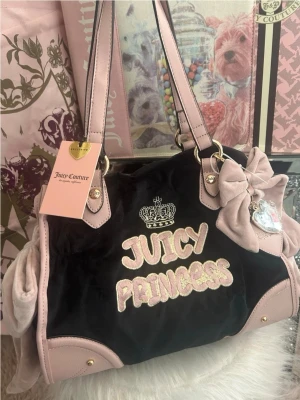 Juicy Couture Juicy Princess handväska - Svart och ljusrosa handväska i sammet från Juicy Couture med broderad text 'Juicy Princess' och krona framtill. Väskan har rosa detaljer, dekorativ rosett och hjärtberlock i metall samt dubbla axelband. Perfekt för dig som gillar en söt och trendig look.