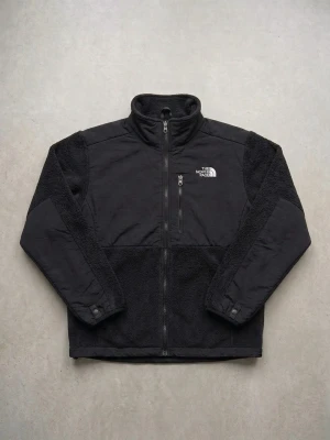 Svart fleecejacka från The North Face - Svart fleecejacka från The North Face med hel dragkedja, hög krage och broderad logga på bröstet och ryggen. Jackan har mjuk, fluffig fleece och släta paneler på axlar och ärmar. Perfekt för kyliga dagar.