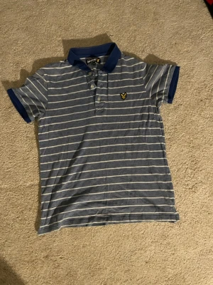 Blå randig piké från Lyle & Scott - Snygg blå pikétröja från Lyle & Scott med vita horisontella ränder och klassisk krage. Tröjan har korta ärmar och knäppning framtill samt den ikoniska gula loggan på bröstet. Perfekt till jeans eller shorts för en avslappnad look.