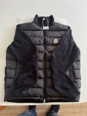 Svart dunväst från Moncler - Snygg svart dunväst från Moncler med stickade ärmar och ribbade muddar. Västen har dragkedja framtill och Moncler-logga på bröstet. Perfekt för lager-på-lager och ger en clean look till din outfit.#grisch #2000s #y2k #archive #niche #2016s #fakemink #feng #youngthug #stockholm #ungdom #snygg #marinblå #svart #vit #fin #unik #123 #456 #789 #archivedesigner #timelessdesign #vintage #vinted #fashion #collectorsitem #vintagedesigner #mönster #man #boy #afflction #replay #gats #maisonmargiela #faded 