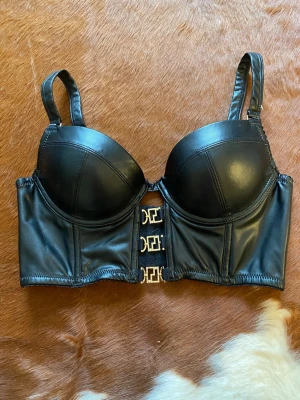Svart bustier-bh i fejkläder - Snygg svart bustier-bh i fejkläder med vadderade kupor och bredare band. Framtill har den tre dekorativa guldfärgade spännen som stängning. Materialet är glansigt och ger en edgy look. Perfekt för dig som vill ha något extra till outfiten. Aldrig använd
