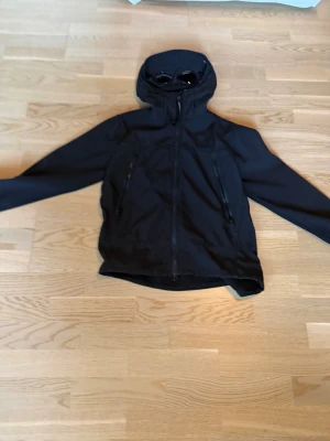 Svart vindjacka från C.P. Company - Säljer en svart jacka från C.P. Company med ikoniska glasögon i huvan. Jackan har dragkedja framtill, två sidofickor med dragkedja och en sportig look. Materialet är slätt och vindtätt, perfekt för blåsiga dagar.